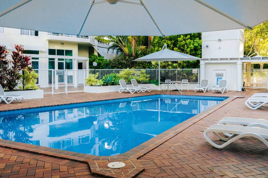Pool with large grey umbrella, white loungers and apartment buildings in background