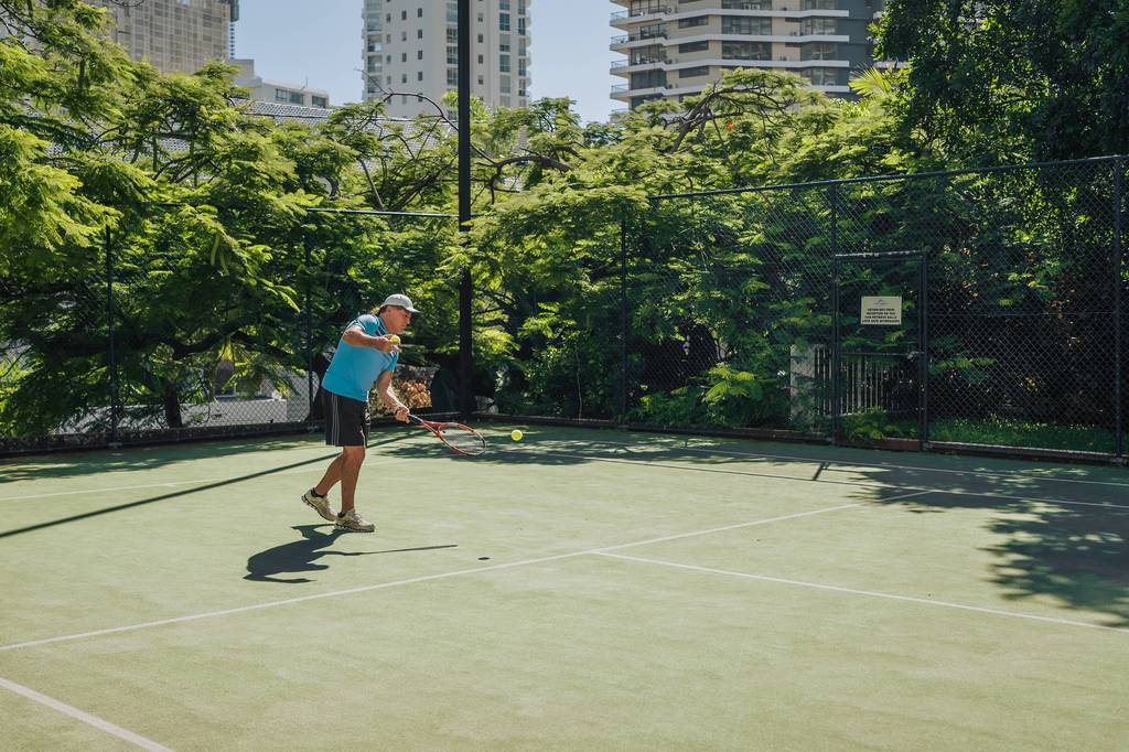 Player serves on a lush green court with shaded tree canopy overhead